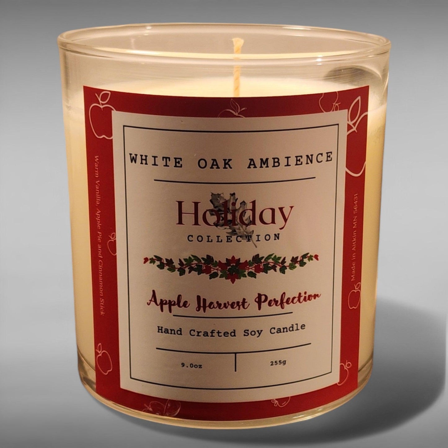 A clear glass jar soy candle with a red and white label reading 'Holiday Collection Apple Harvest Perfection Hand Crafted Soy Candle'. It features a single lit wick in the center.