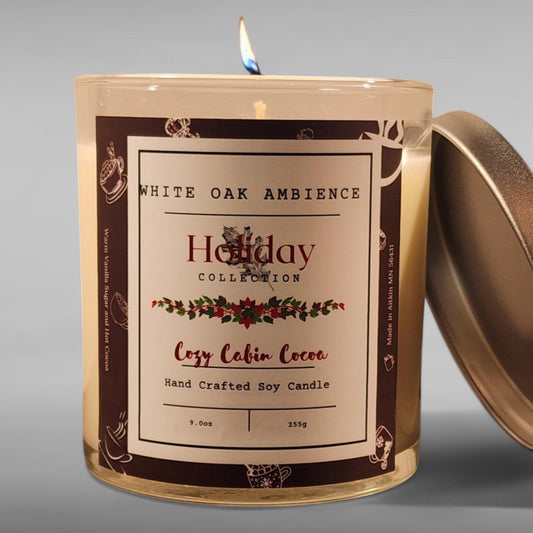 A Cocoa scented, lit soy candle from White Oak Ambience's Holiday collection, with a label that reads 'Cozy Cabin Cocoa'. The candle is in a clear container with a brown label, placed on a grey surface.