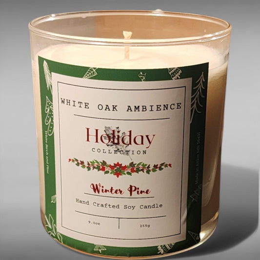 A clear glass jar containing a white soy candle with a green and white label reading 'Winter Pine Hand Crafted Soy Candle', with holiday decorations depicted.